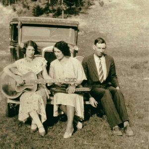 The Carter Family trio sit on the front bumper of car in a field. 