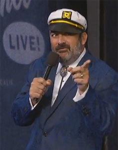 Jonathan Kimmel, a burly bearded man, dances with a microphone in a jaunty sailor cap and dark blue suit jacket.