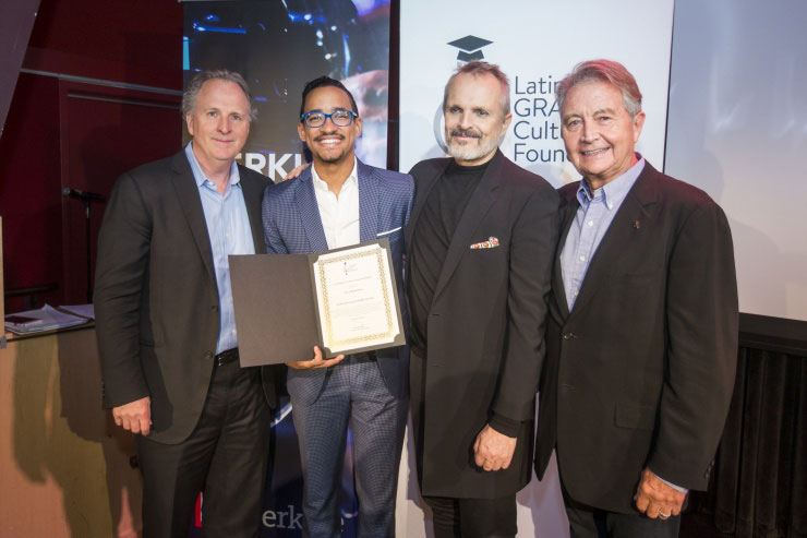 Four suited, smiling gentlemen face the camera, one holding a scholarship