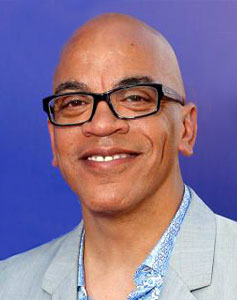 In a light-gray sport jacket, blue shirt and black-framed glasses, Rickey Minor smiles for the camera.