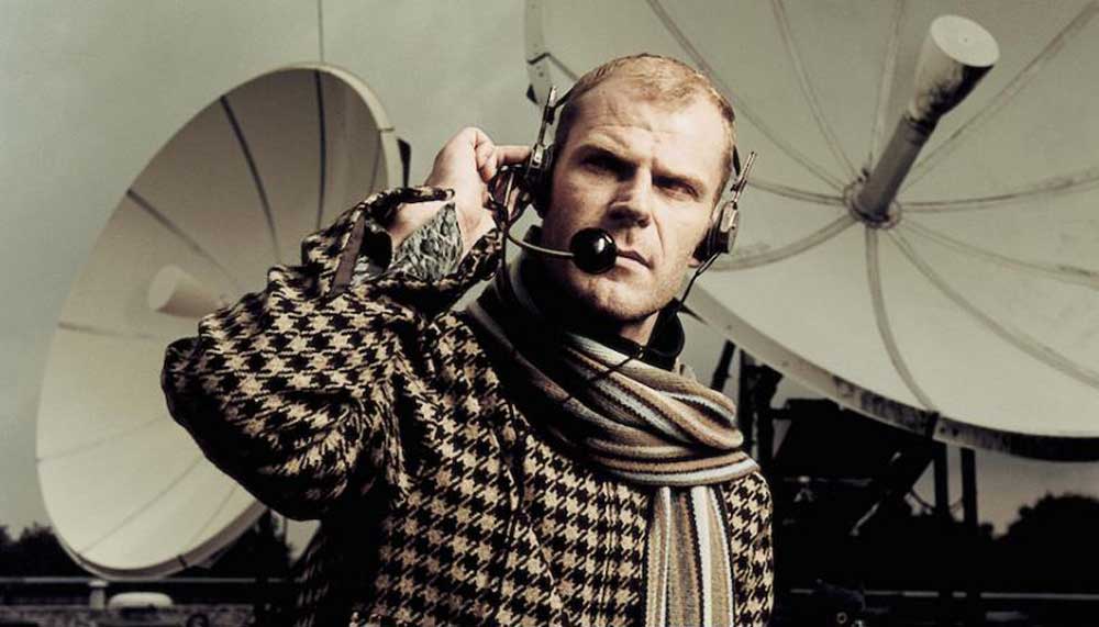 Tom Holkenborg poses in a houndstooth checked jacket with a headset, in front of satellite dishes.