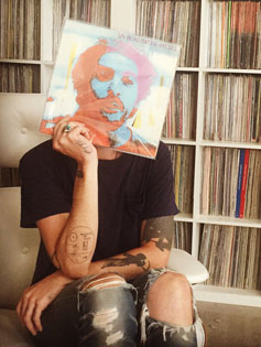 Zach Cowie sits in front of a wall of records, holding a vinyl LP with the an image of a face on it in front of his own face, becoming one with the music.