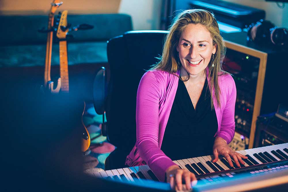 Ronit Kirchman at the keyboard in a pink sweater, with guitars in background.