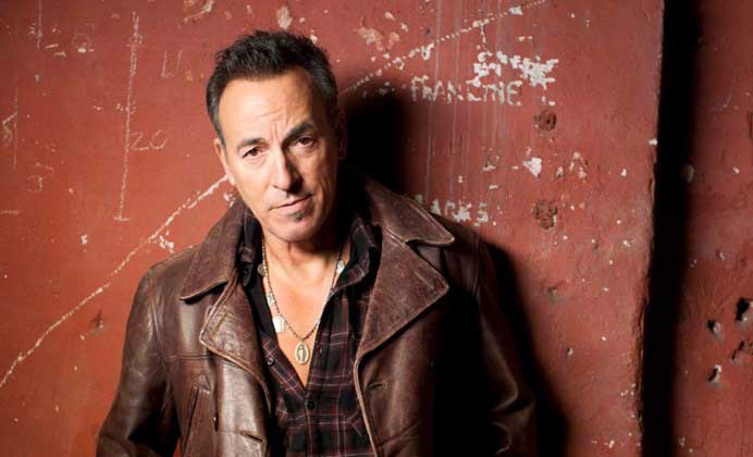 Bruce Springsteen in a brown leather jacket against a terra cotta colored wall. 