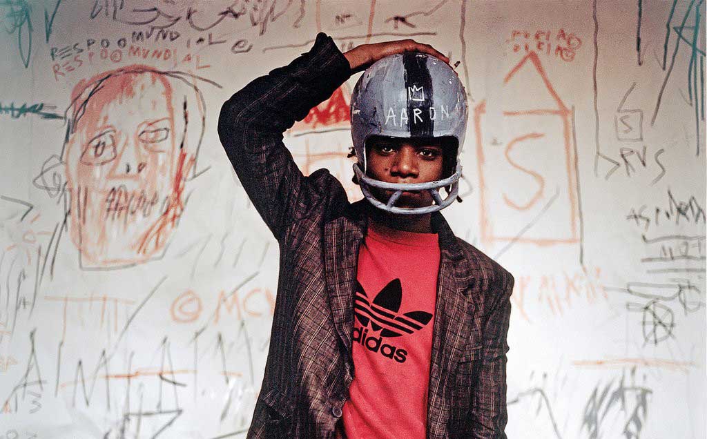 Jean-Michele Basquiat in a football helmet stands in front of one of his paintings.