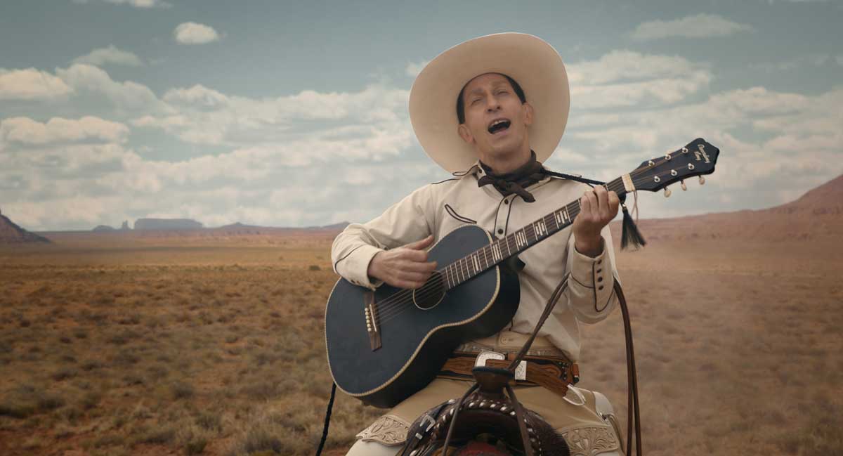 Tim Blake Nelson belts out a song on acoustic guitar wearing a cream-colored cowboy hat.