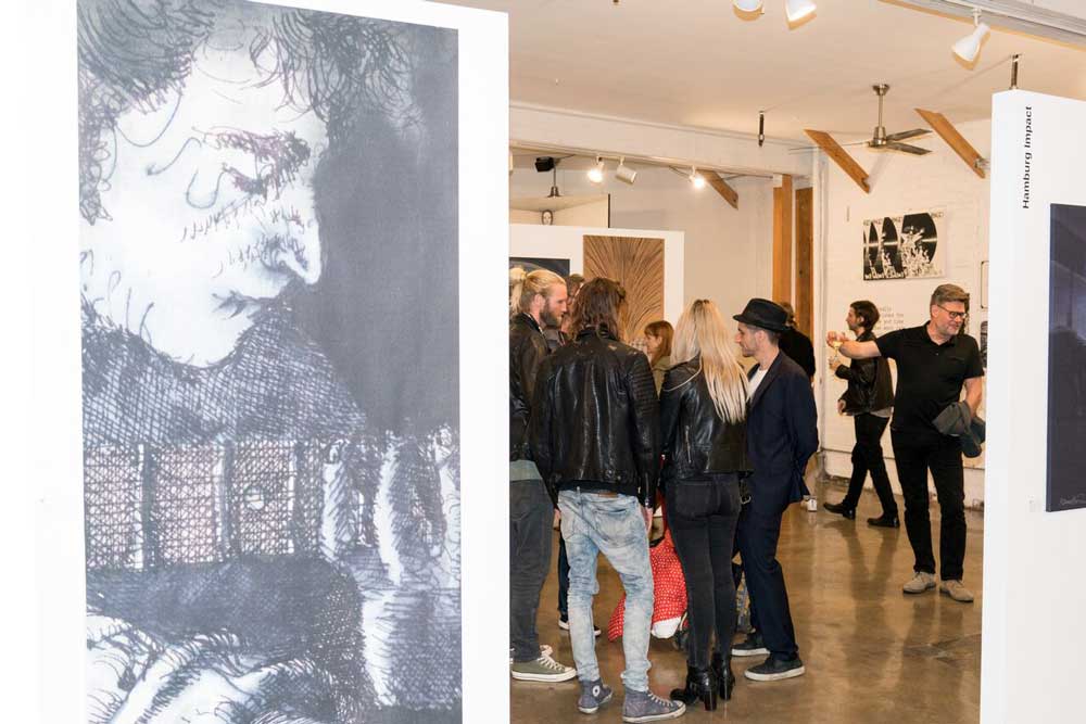 Kalus Voormann line-drawing of John Lennon hangs in foreground as people socialize in the gallery.