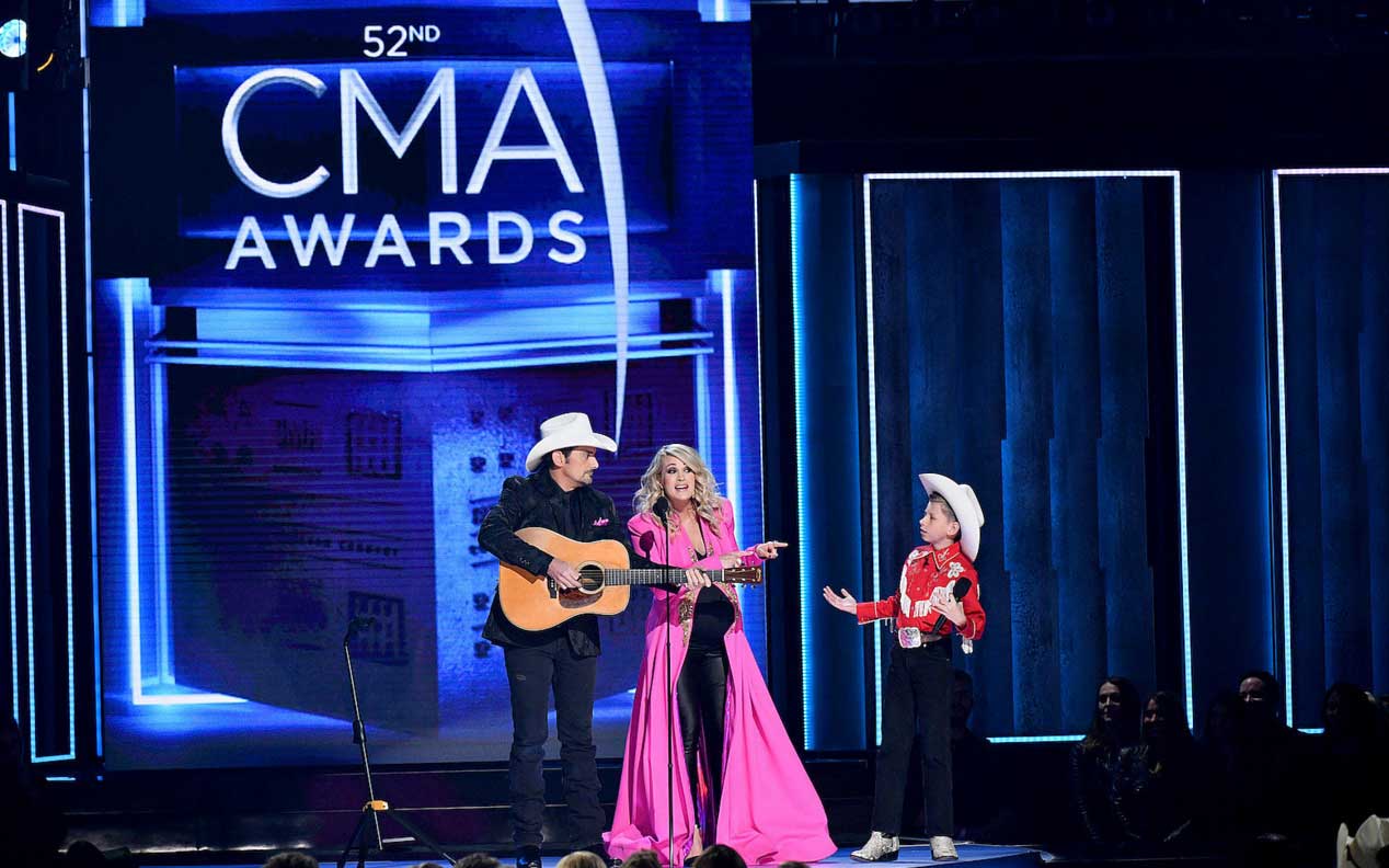 Brad Paisley strums an acoustic guitar while pink be-gowned Underwood and kid in cowboy hat look on.