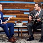 Chris Isaak strums a guitar as Dave Karger gestures with a sheaf of papers.